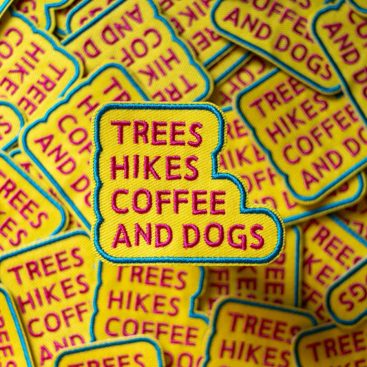 A Patch with 'Trees Hikes Coffee and Dogs' text on a yellow background from the Pencil Me In stationery shop.
